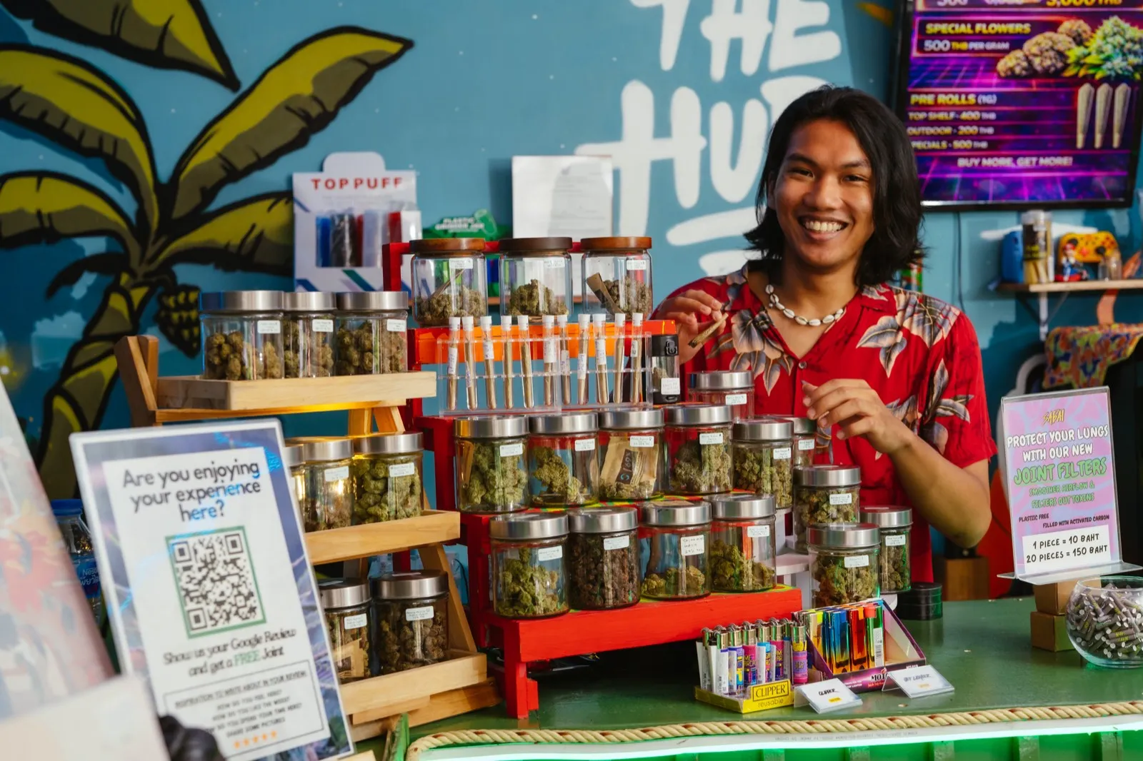 Smiling staff at The Hub's cannabis counter, lined with jars of flower beneath the mural, Ao Nang